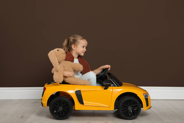 Cute little girl with toy bunny driving children's car near brown wall indoors - Φωτογραφία, εικόνα