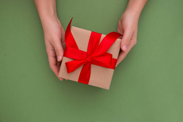 Female hands are holding a gift box on a green background, copy space. Gifts and presents concepts. View from above. - Photo, Image