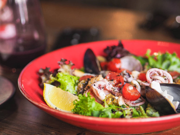 Fresh mediterranean seafood salad with olive oil, lemon, tomatoes and lettuce in red plate on the table in restaurant - Фото, изображение