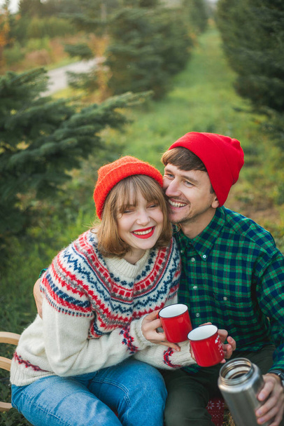 Christmas. Happy young couple in love in red hats and sweaters drink hot tea from thermos near Christmas trees. Christmas tree market. - Fotografie, Obrázek