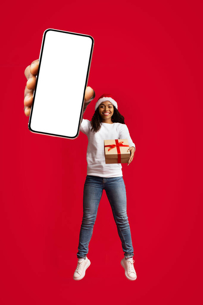 Ευτυχισμένη μαύρη γυναίκα σε Σάντα καπέλο κρατώντας smartphone, δώρο Χριστούγεννα - Φωτογραφία, εικόνα
