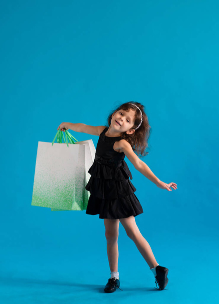 Cute cheerful girl in black dress with paper bags for shopping on blue background - Фото, зображення