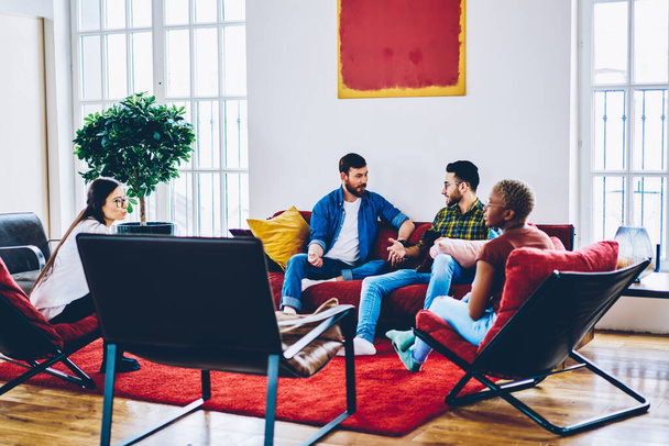 Young group of creative male and female colleagues discussing design ideas in modern interior with red furniture, teen multiracial hipsters spending leisure time for talking during guests meeting - Valokuva, kuva