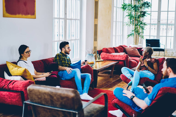 Young multicultural hipster guys in casual clothes talking about friendship and socialising during fellow meeting in modern living room, diverse guests discussing trendy decoration in home flat - Фото, зображення