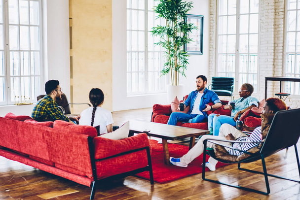 Good looking hipster guys in stylish outfits enjoying weekend time in lounge zone of loft apartment, diverse male and female colleagues with takeaway coffee to go communicate about recreation - Fotoğraf, Görsel