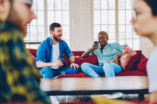 Young hipster guys in trendy clothes relaxing togetherness in loft apartment, multiracial male and female best friends with takeaway cups discussing fellowship during coffee time in home interior - Photo, Image