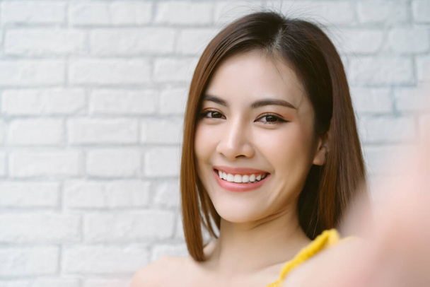Closeup of beautiful Asian woman wearing off shoulder yellow knitted clothes smilling with white teeth and taking selfie of by camera on white brick wall background. Natural Beauty, Healthy Woman. - Fotó, kép
