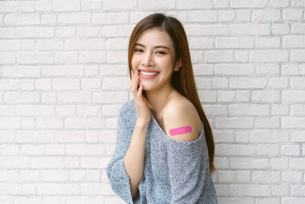 Smiling beautiful Asian woman wearing off shoulder sweater sleeve showing her arm with adhesive bandage plaster after receiving the Covid-19 Vaccine on white brick wall background. Vaccine Safe lives. - Foto, Bild