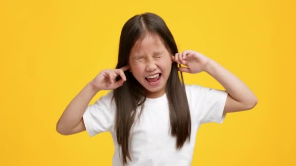 ondeugend klein aziatisch meisje pluggen oren met vingers, gele achtergrond - Video