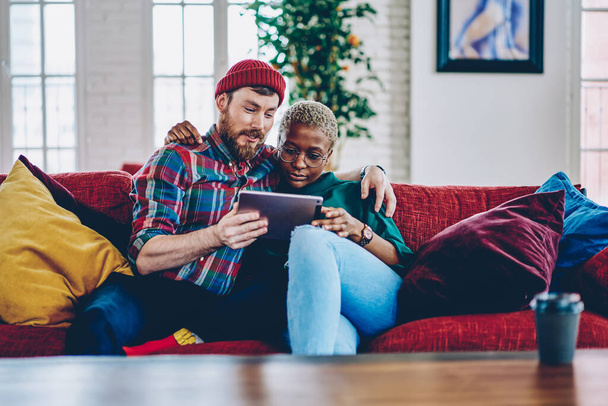 Diverse hipster guys hug and reading received email message using home internet connection on modern touch pad, Caucaisan husband and African American wife in eyewear browsing wireless website - Foto, imagen