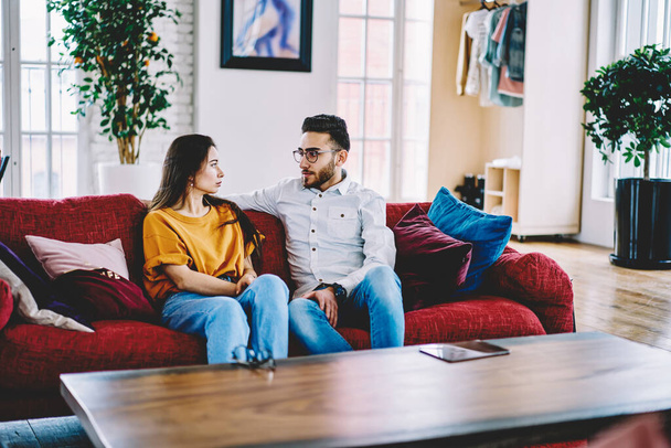 Diverse hipster guys in casual apparel communicate about friendship resting on comfortable red furniture in apartment, Turkish husband in optical eyeglasses and Caucasian wife talking about relations - Φωτογραφία, εικόνα