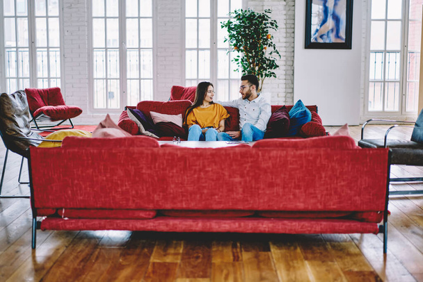 Youthful couple in love talking and discussing plans during weeknd time in apartment, Turkish boyfriend communicating with Caucasian girlfriend resting at cozy sofa in stylish living room - Фото, изображение