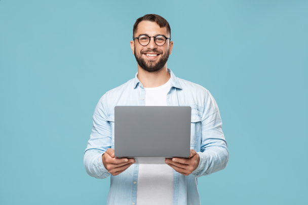Vrolijke volwassen Europese man in bril met laptop kijkt naar camera, geïsoleerd op blauwe achtergrond - Foto, afbeelding