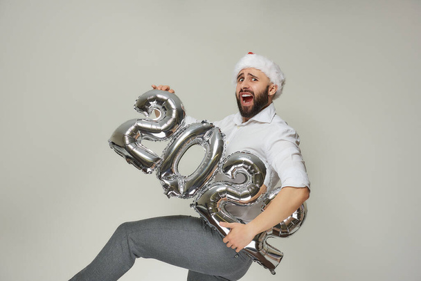 Een verwarde man met een baard in een rood fluwelen kerstmuts houdt zilveren ballonnen in de vorm van 2022 en tilt zijn been op en schreeuwt. Nieuwjaarsfeest. - Foto, afbeelding