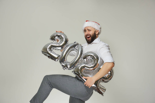 Zmatený muž s vousy v červeném sametovém klobouku Santa drží stříbrné balónky ve tvaru roku 2022, zvedá nohu a křičí. Novoroční párty. - Fotografie, Obrázek