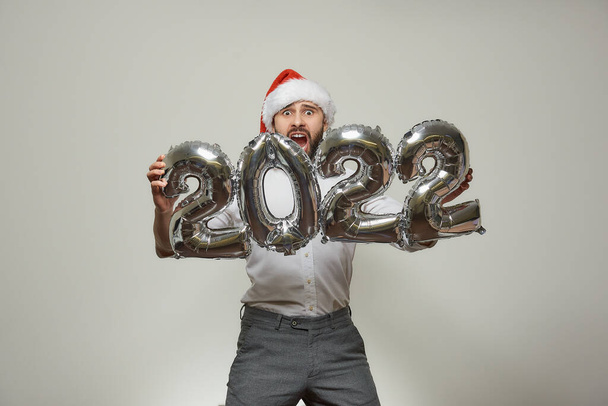 Een ongelukkige man met een rode fluwelen kerstmuts houdt zilveren ballonnen in de vorm van 2022 en schreeuwt van angst. Een man met een baard op een nieuwjaarsfeest. - Foto, afbeelding