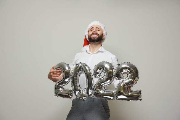 Dumny człowiek w czerwonym aksamitnym kapeluszu Mikołaja trzyma srebrne balony w kształcie z 2022 roku. Szczęśliwy facet z brodą na imprezie noworocznej. - Zdjęcie, obraz