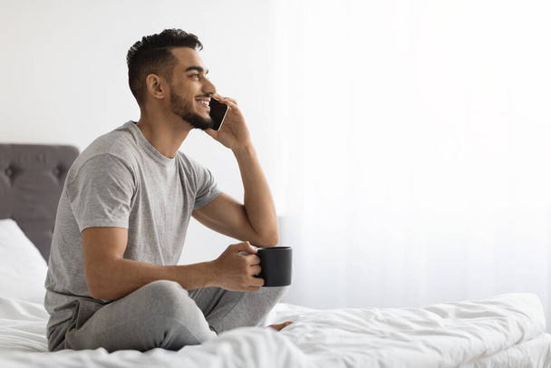 ベッドでリラックスしながら携帯電話で話をし、コーヒーを飲むアラブ人男性 - 写真・画像