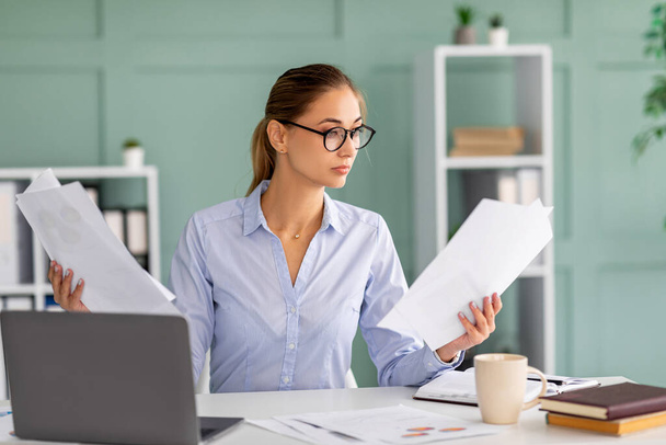 Konzentrierte Geschäftsfrau, die am Laptop arbeitet und Berichte hält, am Arbeitsplatz im Bürointerieur sitzt und Dokumente liest - Foto, Bild