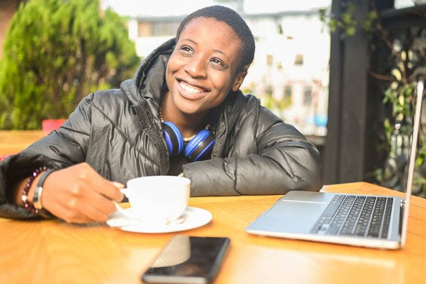 Sorrindo feliz Africano preto mulher de cabelos curtos estudante em preto jaqueta com fones de ouvido azuis tomando café no café da rua trabalhando no computador portátil. Diversidade. Trabalho à distância, educação à distância. - Foto, Imagem
