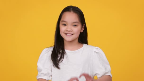 Szeretet és könyörület. Aranyos kis ázsiai lány mutató szív gesztus és mosolygó kamera, kifejező kedvesség - Felvétel, videó