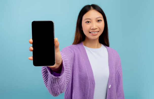 Cool aplicación o sitio web. Señora asiática mostrando teléfono inteligente con pantalla vacía, de pie sobre fondo de estudio azul, primer plano - Foto, Imagen