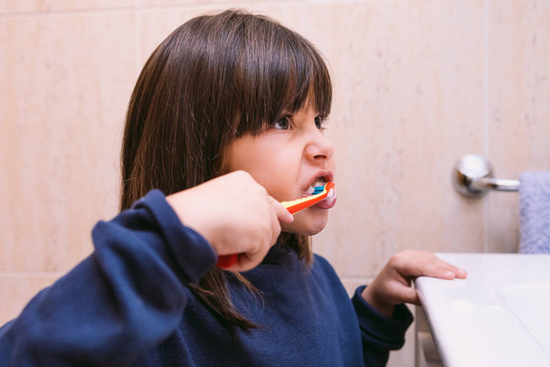 Pieni tyttö tummansininen huppari, harjaten hampaitaan katsoessaan peiliin, kylpyhuoneessa. Hammasharjaus, hygienia ja lapsuuden käsite. - Valokuva, kuva