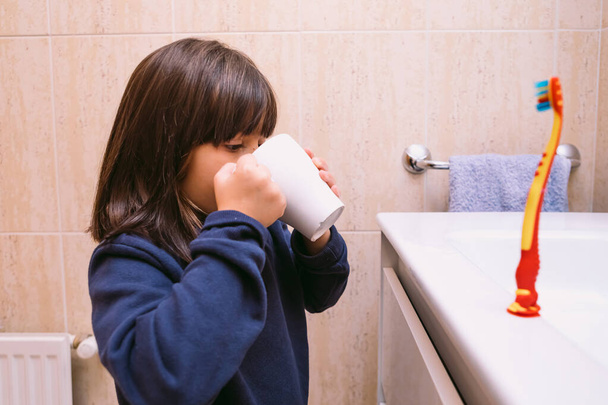 Petite fille en sweat-shirt bleu foncé, se brossant les dents, se rinçant la bouche avec de l'eau dans l'évier, dans la salle de bain. Brossage des dents, hygiène et concept d'enfance. - Photo, image