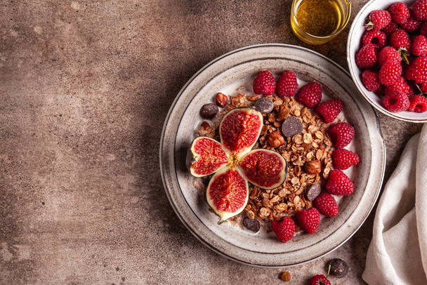 Chocolate granola with almond milk, raspberries, figs and honey in a ceramic plate on a brown background. Healthy breakfast or snack. - Photo, image