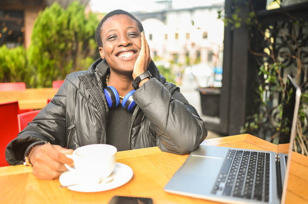 Smiling happy African black short-haired woman student in black down jacket with blue headphones having coffee at street cafe working on laptop computer. Diversity. Remote work, distance education. - Photo, Image