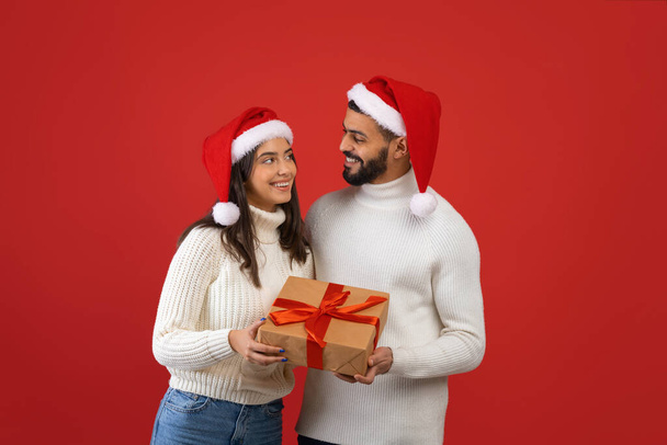 Güzel Arap çift Noel hediyelerini gösteriyor ve birbirlerine bakıyorlar, kırmızı arka planda duruyorlar, boş yer var. - Fotoğraf, Görsel