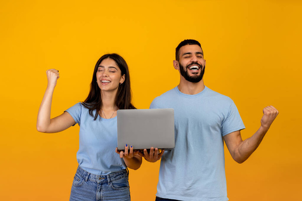 Big-Win-Konzept. Glückliches arabisches Paar hält Laptop in der Hand, jubelt und hebt die Hände, feiert den Sieg, gelber Hintergrund - Foto, Bild