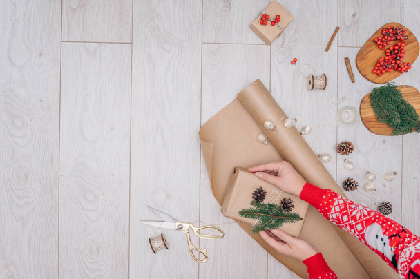 女性の手は茶色の紙にクリスマスや休日の手作りプレゼントを包む。白い木製のテーブルの上のボックスと装飾、コピースペース付きのトップビューを提示します。DIY趣味. - 写真・画像