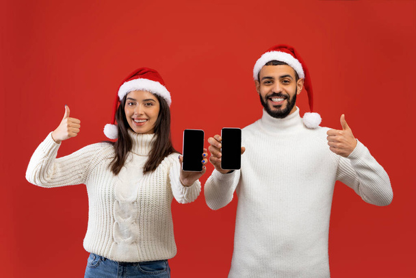 Coppia araba in cappelli Babbo Natale che mostra schermi vuoti per la pubblicità natalizia, gesticolando pollici in su - Foto, immagini