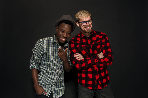 Studio shot of two stylish young men having fun. Handsome bearded hipster in a shirt in a cage standing next to his African-American friend in hat against a dark background. - Foto, imagen