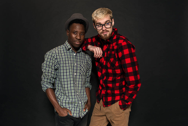 Studio ampui kaksi tyylikästä nuorta miestä hauskaa. Komea parrakas hipsteri paidassa häkissä seisoo afroamerikkalaisen ystävänsä vieressä hatussa tummaa taustaa vasten.. - Valokuva, kuva