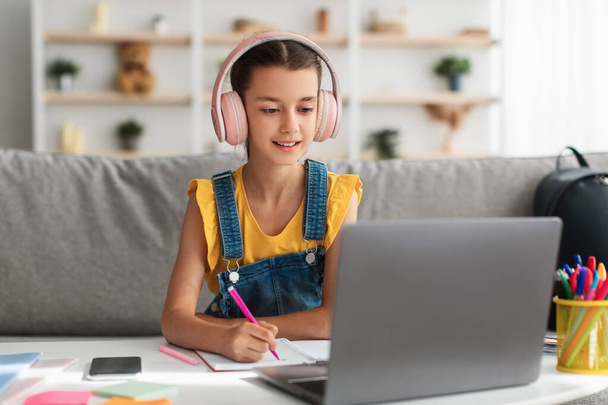 Schoolgirl in wireless headset sitting at desk, writing in textbook - Foto, immagini