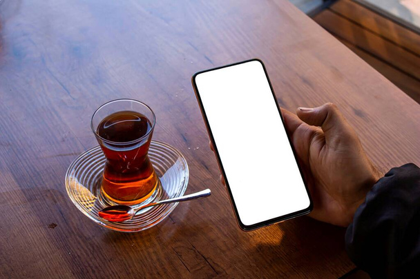 Άνθρωπος που χρησιμοποιεί smartphone, mockup εικόνα ενός άνδρα χέρι που κρατά το κινητό τηλέφωνο με λευκή οθόνη ενώ κάθεται σε ένα καφέ. - Φωτογραφία, εικόνα