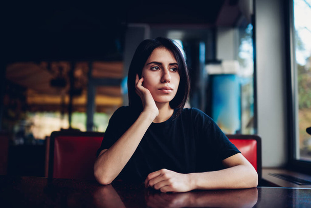 Young dreamy ethnic female in casual outfit with dark hair looking away while sitting at table near window in cafe leaning on hand - Photo, Image