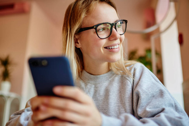 Счастливая девушка-хипстер в классических очках, отводящая взгляд и улыбающаяся в дневное время суток за использование мобильных технологий, миллениалка в экипировке, наслаждающаяся отдыхом для сетевого общения - Фото, изображение