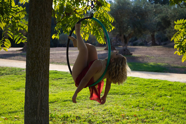 Mujer rubia y joven atleta acrobat gimnasta realizando ejercicio aéreo en el aire libre anillo en el parque. Mujer flexible en traje rojo realiza poses de artistas de circo bailando con caderas. - Foto, Imagen