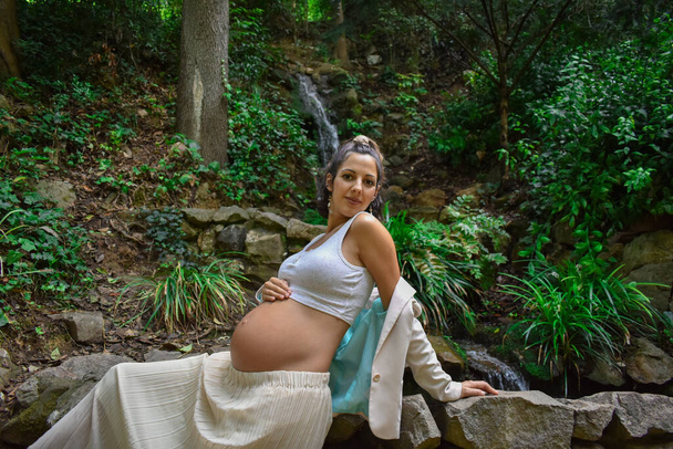 A beautiful young Caucasian pregnant woman in white clothes posing in nature leaning on a stone wall - Fotografie, Obrázek