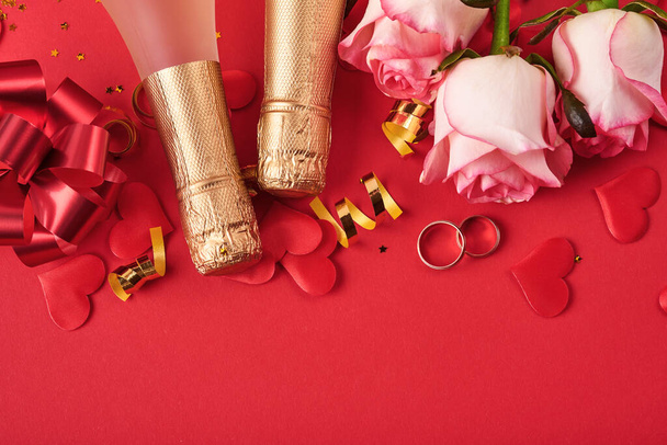 Rosas rosadas flores, anillos de boda, champán, regalo, cintas doradas y corazones de confeti sobre fondo rojo. Vista superior con espacio para saludos. Día de San Valentín o fondo de boda y tarjeta de felicitación. - Foto, Imagen