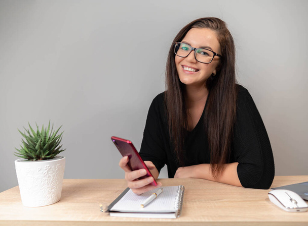 Щаслива жінка використовує мобільний телефон під час роботи вдома з ноутбуком. Усміхнена жінка в окулярах обміну повідомленнями зі смартфоном, перегляд сайту на мобільному телефоні
. - Фото, зображення