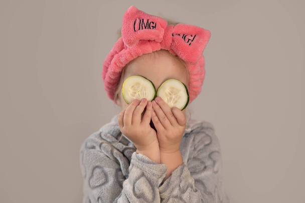Маленька дівчинка в халаті і тримає шматочки огірка на очах
 - Фото, зображення