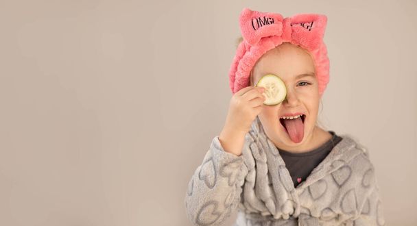 Una bambina che indossa accappatoio e tiene pezzi di cetriolo sugli occhi - Foto, immagini
