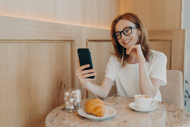 Belle rousse femme dans les écouteurs assis dans le café et faire un appel vidéo sur téléphone mobile - Photo, image