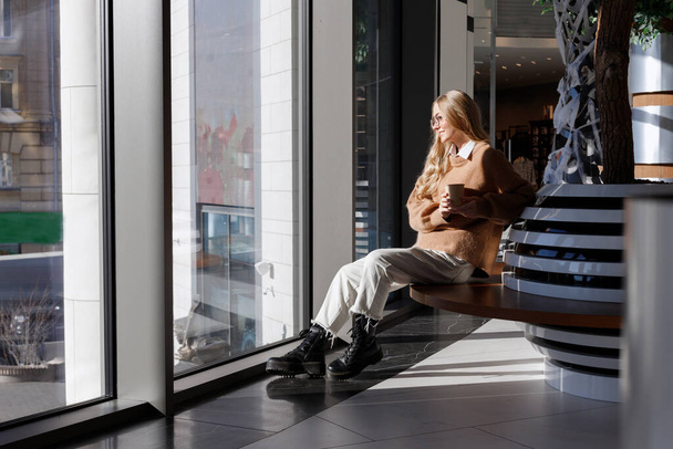 Девушка пьет кофе и смотрит в окно в торговом центре - Фото, изображение
