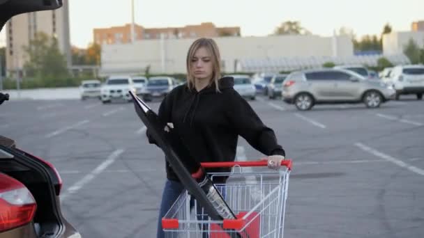 Nainen kuljettaa modernia suurta televisiota supermarketin vaunussa ja yrittää ladata sen auton takakonttiin pysäköintipaikalla. - Materiaali, video