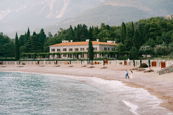 The bride and groom walk holding hands on the pebbled seashore near beautiful villa  - Photo, image